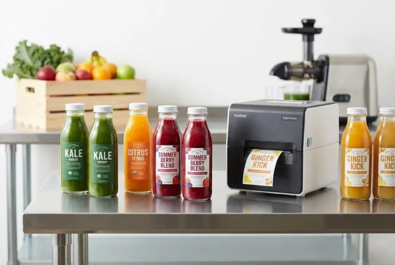 Small Batch Cold Pressed Juice Bottles With Custom Labels Next To A Digital Label Printer For Seasonal Varietals.